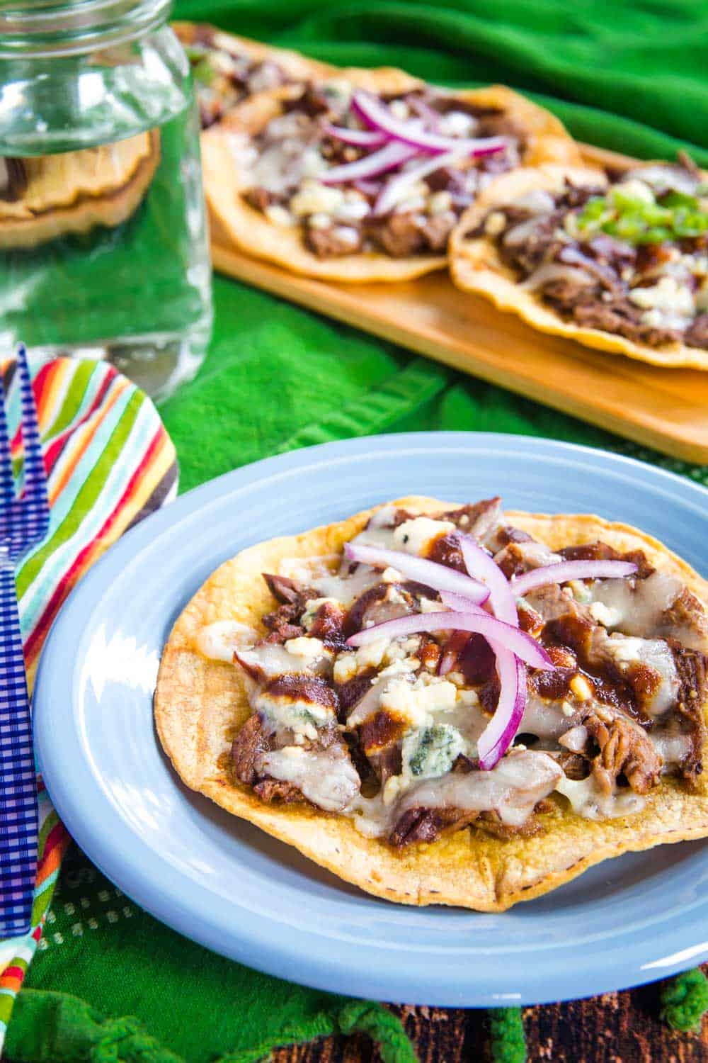 one steak tostada on a blue plate next to a fork and a glass of water with several steak tostadas on a serving platter