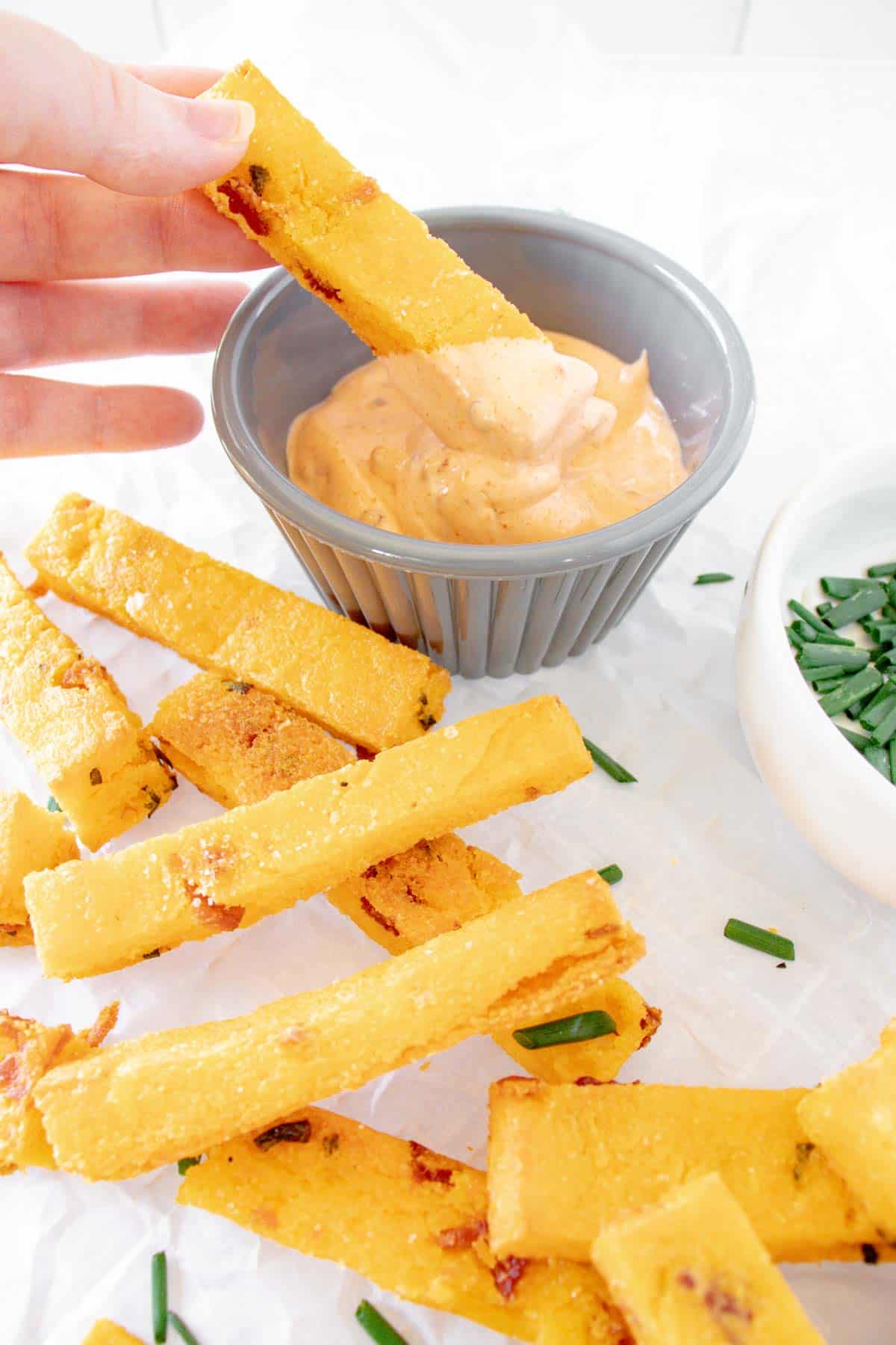 a pile of baked polenta fries and a hand dipping one into a bowl of aioli