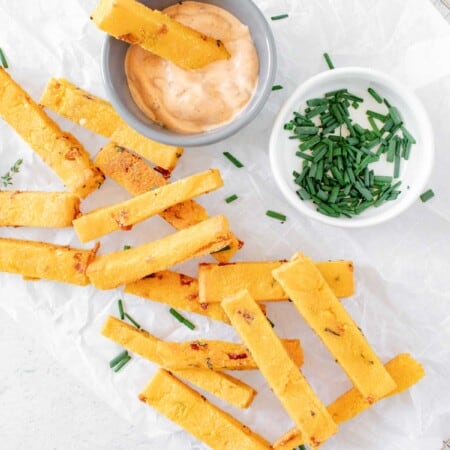 parchment paper on a cooling rack with baked polenta fries laying on it next to bowls of dipping sauce and chopped chives