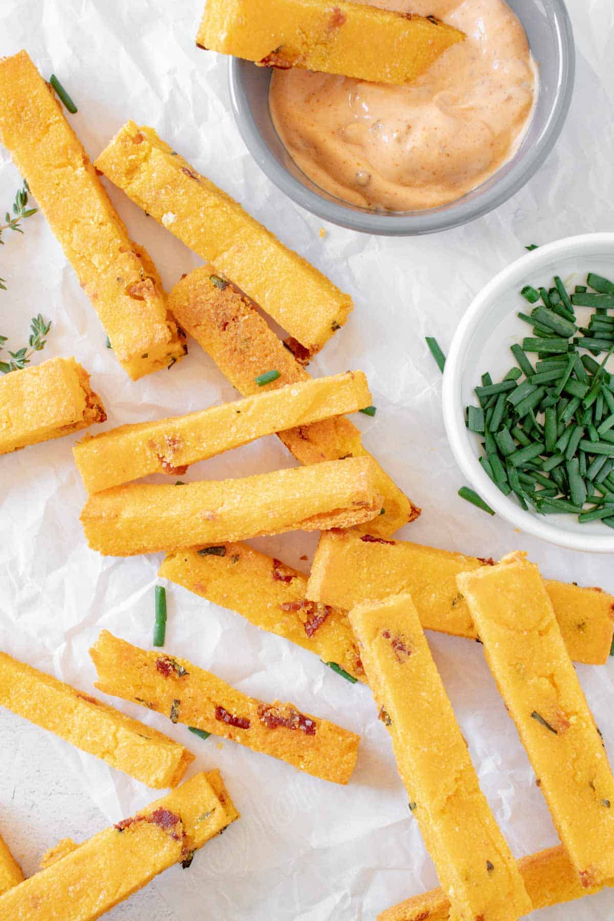 bacon cheddar baked polenta fries on a piece of parchment paper with small bowls of chipotle aioli and minced chives