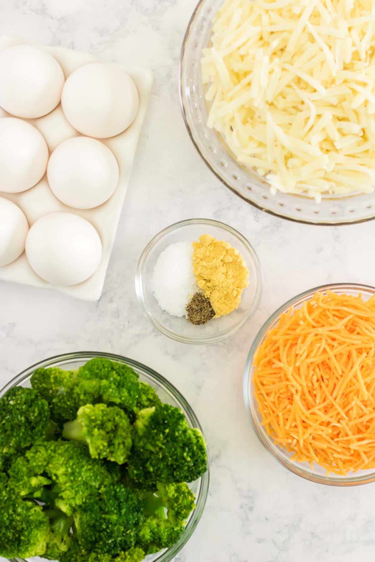 a carton of eggs with bowls of cooked broccoli, shredded cheese, shredded potatoes, and spices on a marble tabletop