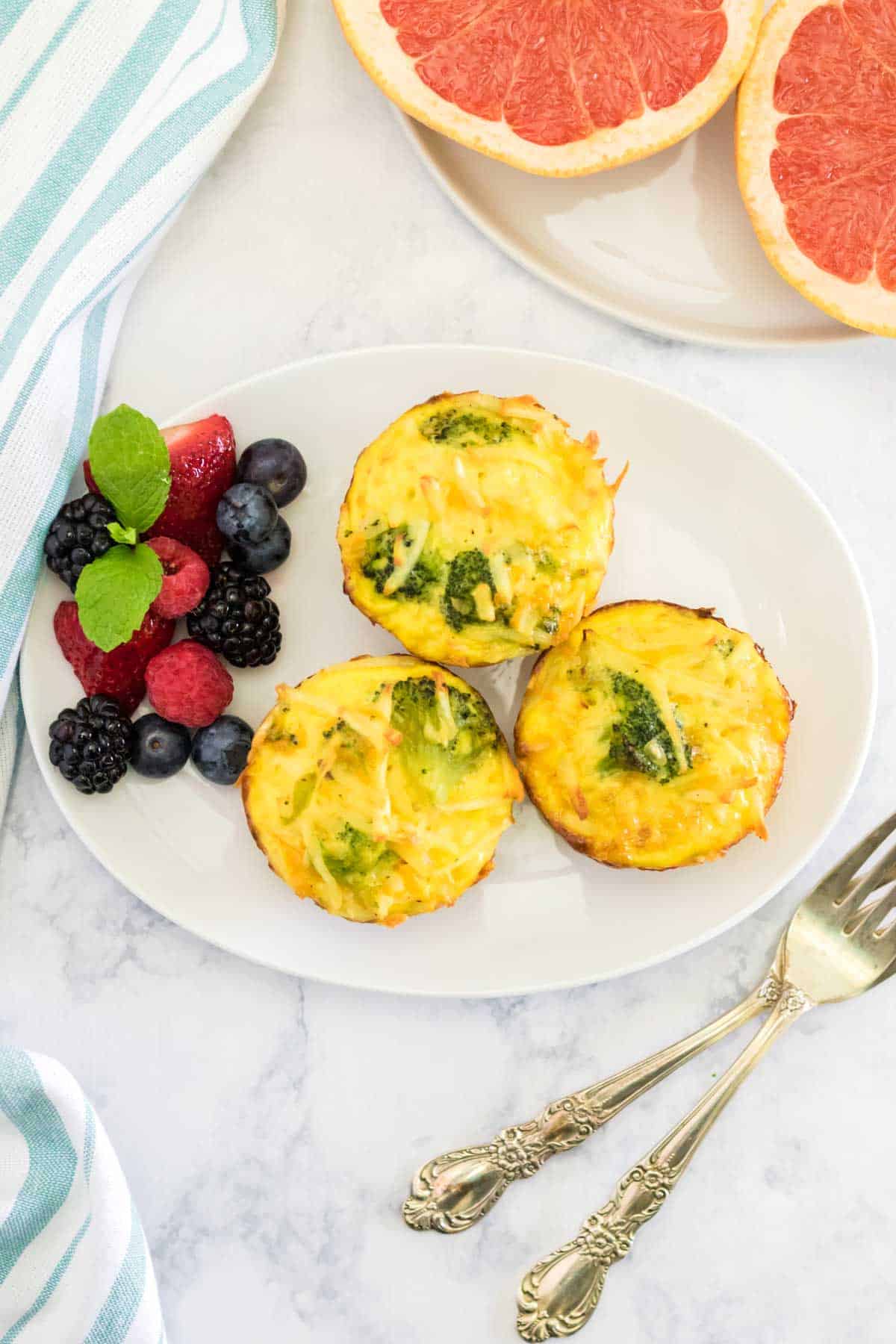 three broccoli egg muffins on a white plate with fruit and a couple of forks on the table