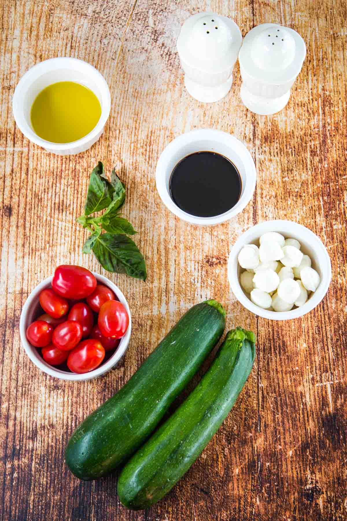 two zucchini, a sprig of basil, salt and pepper shakers, and bowls of grape tomatoes, fresh mozzarella balls, olive oil, and balsamic vinegar on a wooden tabletop