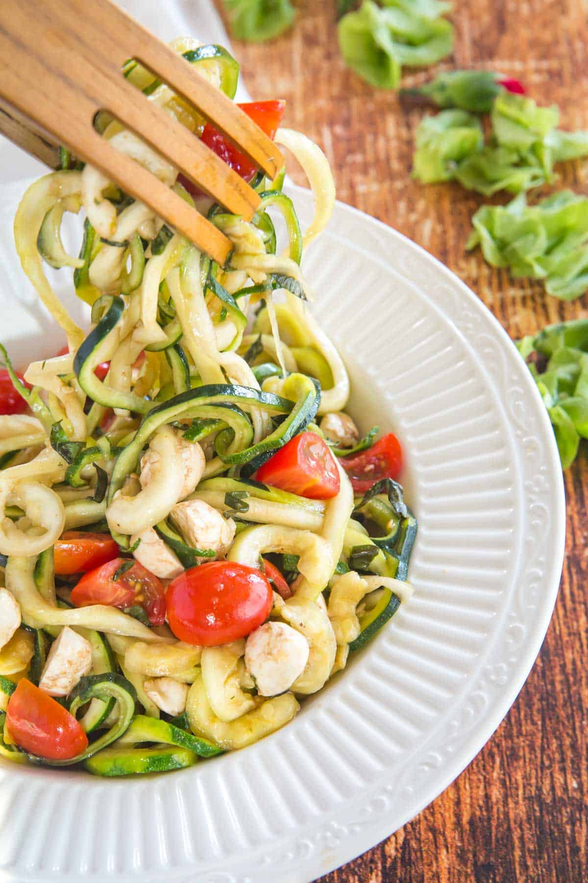 wooden tongs picking up a serving of zoodles with tomatoes and fresh mozzarella