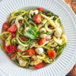 a serving bowl of zoodles with tomatoes, fresh mozzarella, and basil on a wooden table