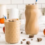 Two pumpkin chocolate smoothies on a counter in front of a white tile wall with white ceramic pitchers of various sizes against the wall