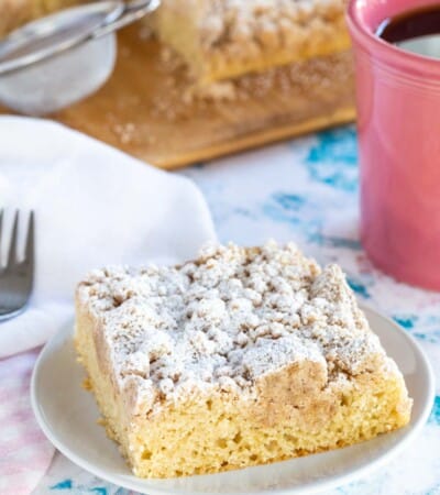 A piece of crumb cake on a white plate with a pink coffee mug near it.