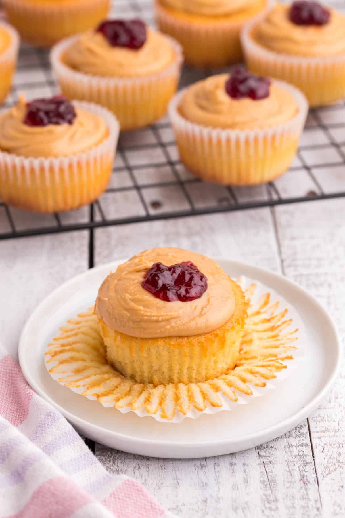 gluten free peanut butter and jelly cupcake on a white plate with the wrapper peeled off and more cupcakes on a rack behind it