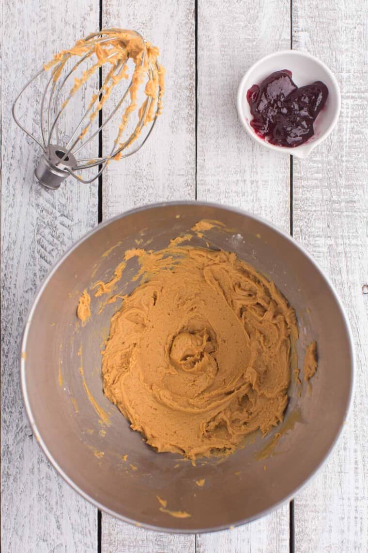 peanut butter frosting in the bowl of a mixer with the whisk attachment next to it and a bowl of jelly