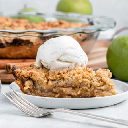 slice of dutch apple pie topped with a scoop of ice cream next to a granny smith apple and the rest of the pie in a glass pie plate