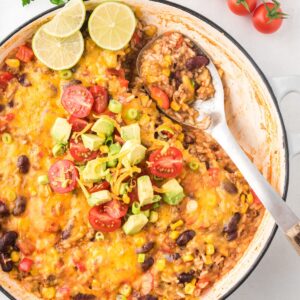 A cheesy taco skillet with rice, beans, and ground beef plus taco toppings piled in the center and a serving spoon in it in the spot where a serving has been removed.