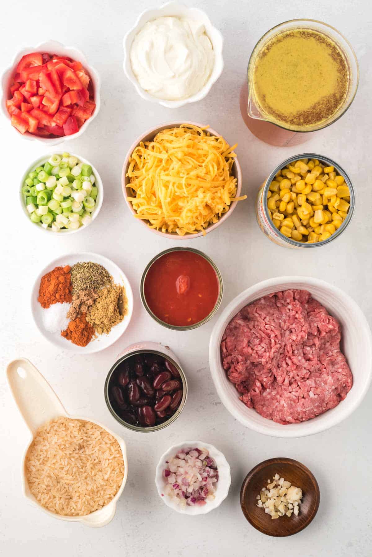 bowls of raw ground beef, garlic, onion, uncooked rice, various spices, sliced green onions, shredded cheesechopped red bell pepper, sour cream, beef broth, and cand of kidney beans, diced tomatoes, and corn on a tabletop