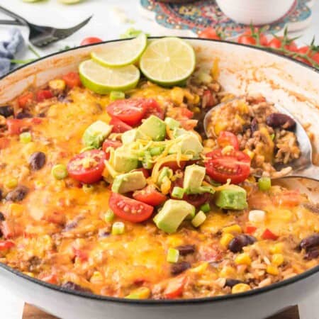 ground beef taco skillet in a pan with serving spoon and a pile of tomatoes, avocado, and scallions on top plus three lime slices