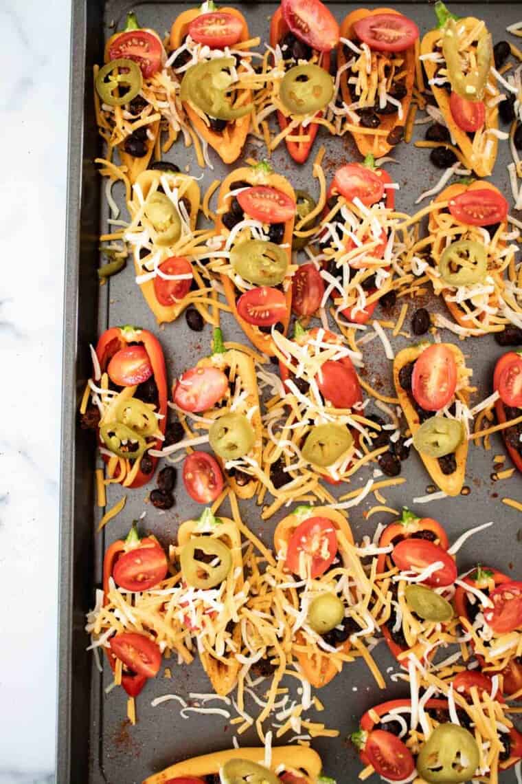 mini bell pepper halves on a sheet pan filled with black beans, grape tomatoes, jalapeno slices, and shredded cheese before baking