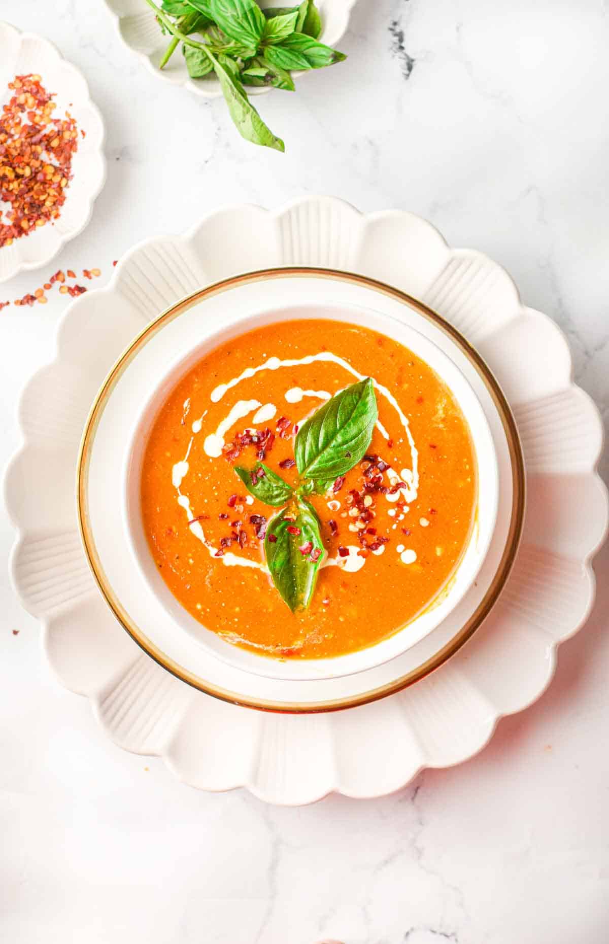creamy tomato soup garnished with a drizzle of cream, basil leaves, and chili flakes in a bowl on top of a sauce with a fluted edge
