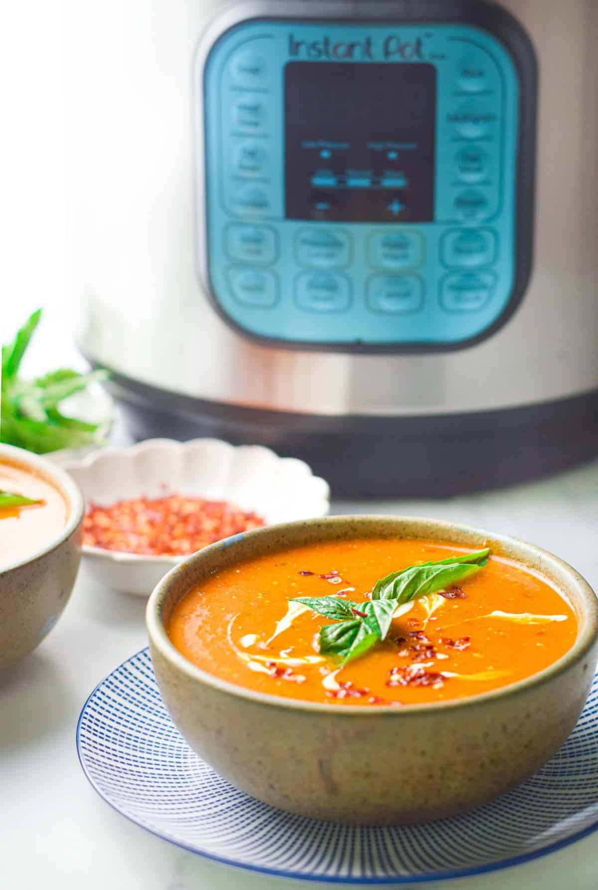 A bowl of tomato basil soup garnished with a drizzle of cream next to a small bowl of chili flakes with the instant pot in the background.