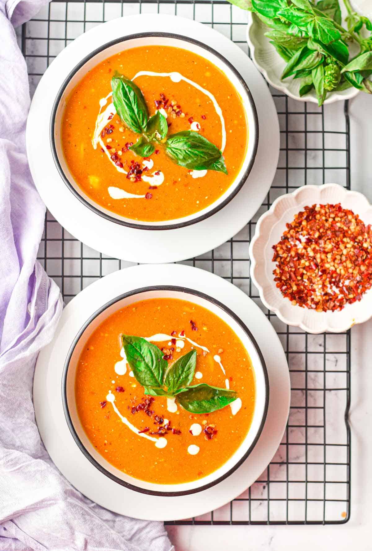 Overhead view of two bowls of creamy tomato soup on a cooling rack next to a bowl of basil leaves and a bowl of red pepper flakes.