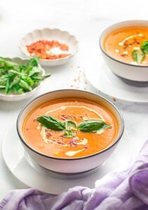Two bowls of instant pot tomato soup garnished with basil and cream next to bowls of basil leaves and chili flakes on a table.