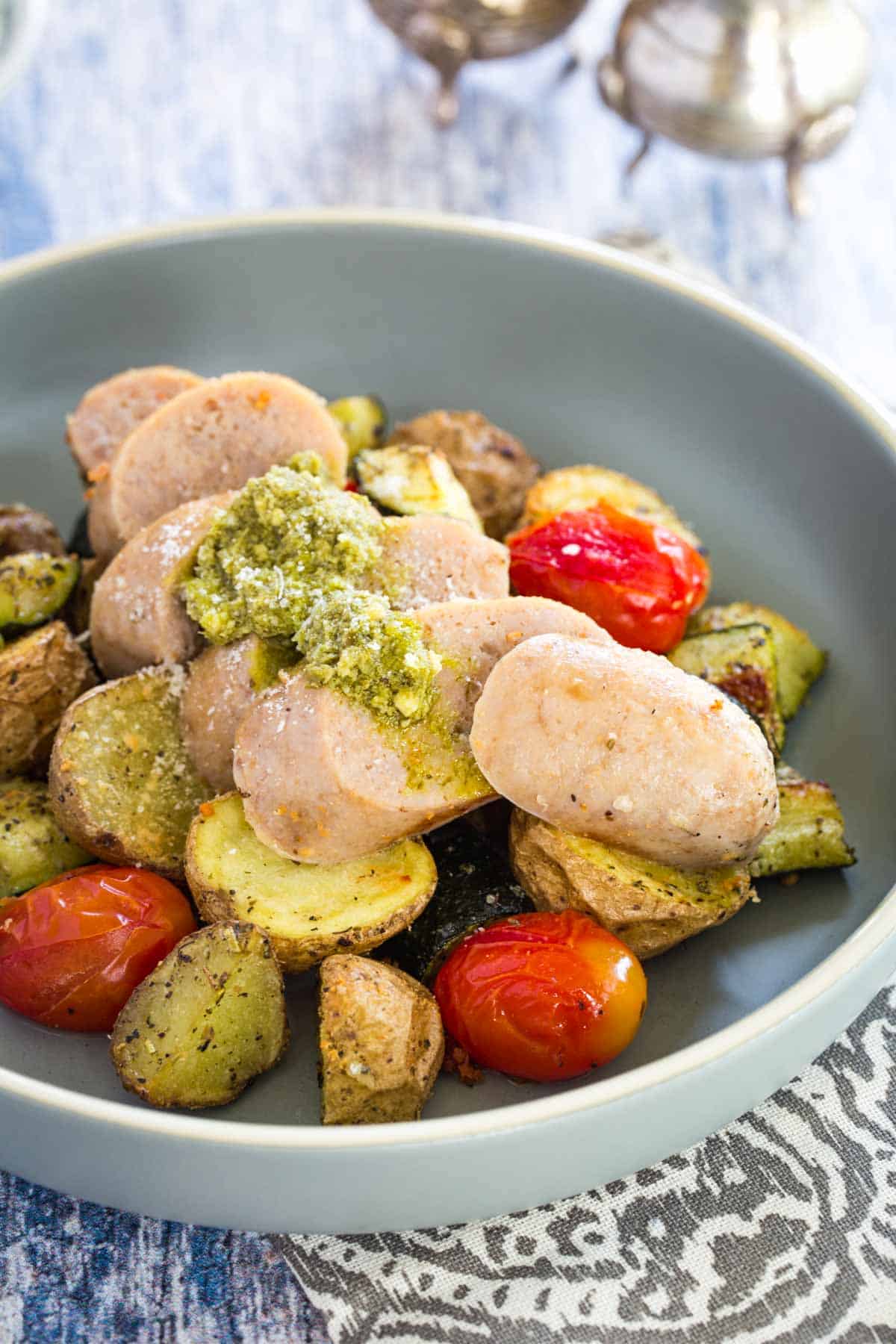 sliced sausage topped with pesto on top of roasted potatoes, zucchini, and tomatoes in a shallow pale blue bowl