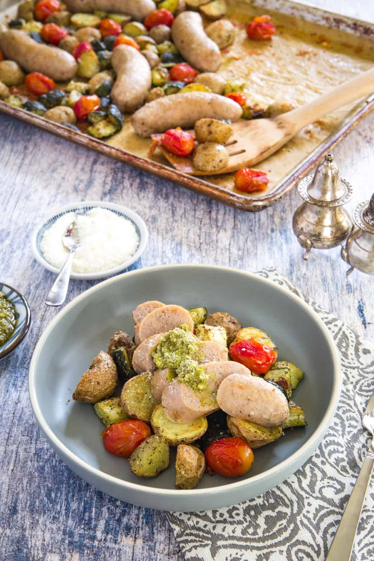 roasted sausage, potatoes, zucchini, and tomatoes in a bowl with the sheet pan dinner behind it on a set table