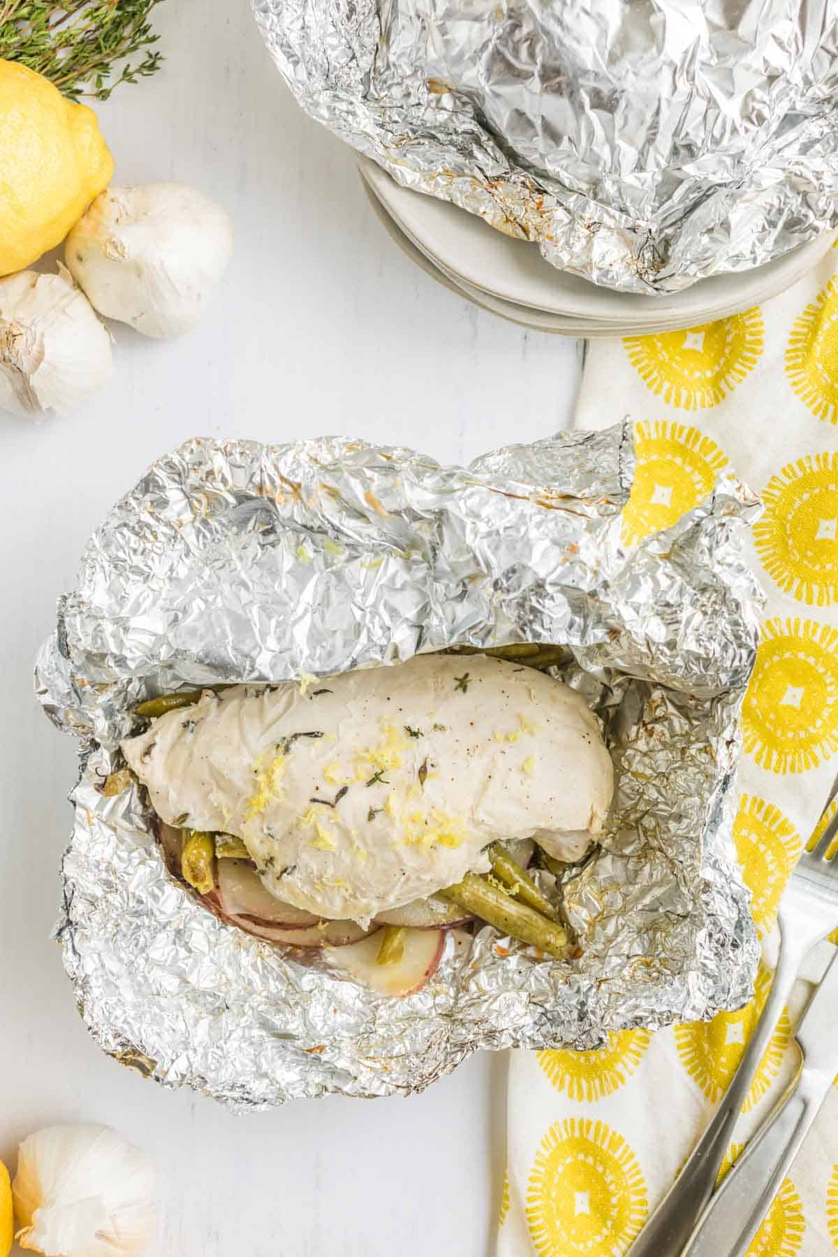 foil pack with chicken breast, green beans, and potatoes next to a yellow and white napkin, lemons, and heads of garlic