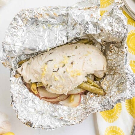 opened up lemon chicken foil packet after it has been cooked next to a yellow and white napkin with a fork and a knife