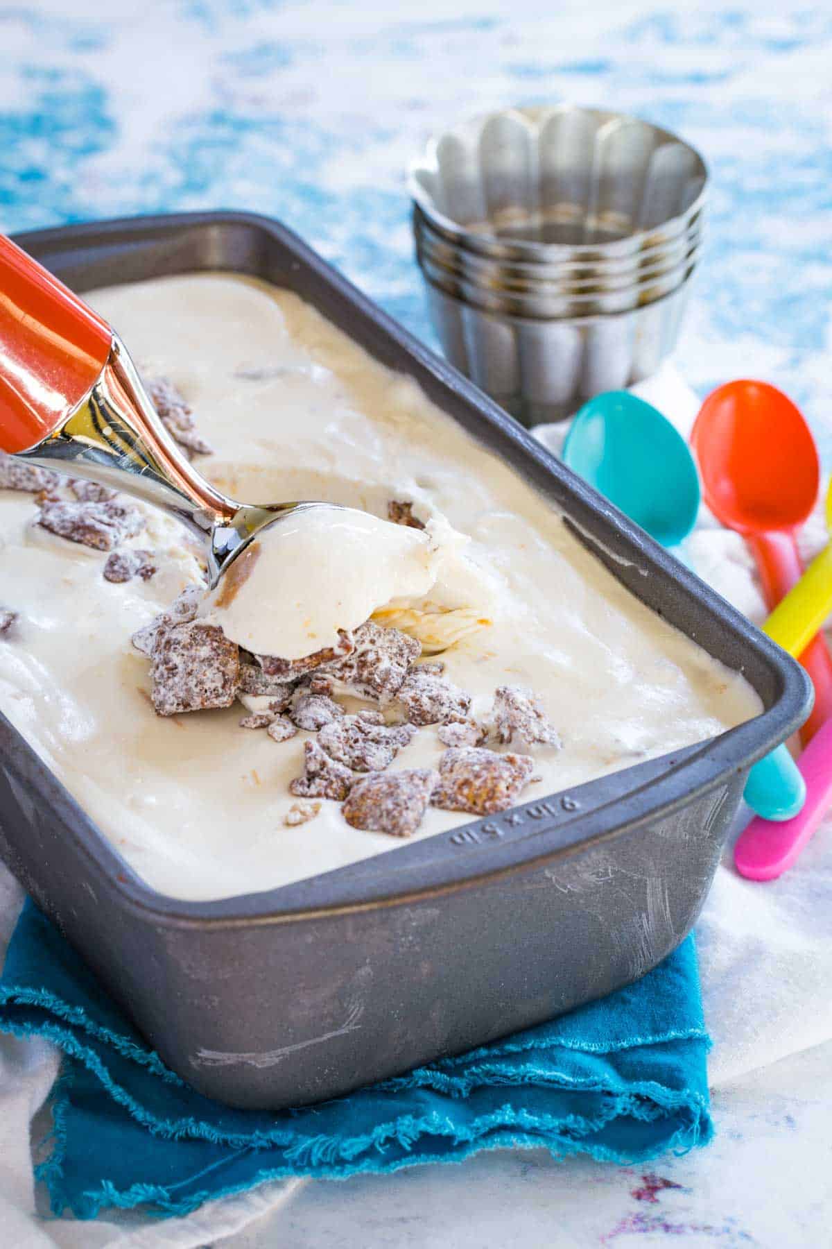 ice cream scoop scooping up muddy buddy ice cream out of a metal loaf pan with small metal cups and multicolored spoons behind it
