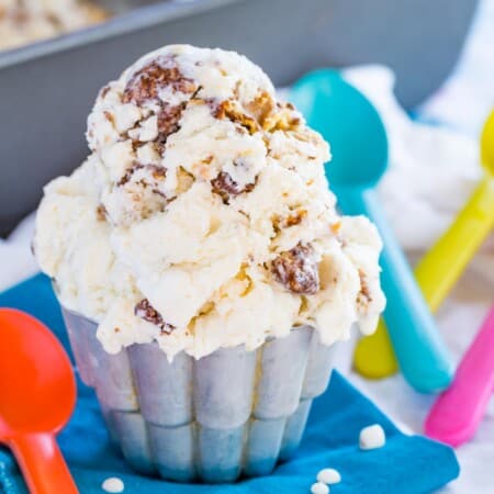 small metal cup of muddy buddy ice cream on top of a blue cloth napkin next to colorful spoons in front of the loaf pan with the rest of the ice cream