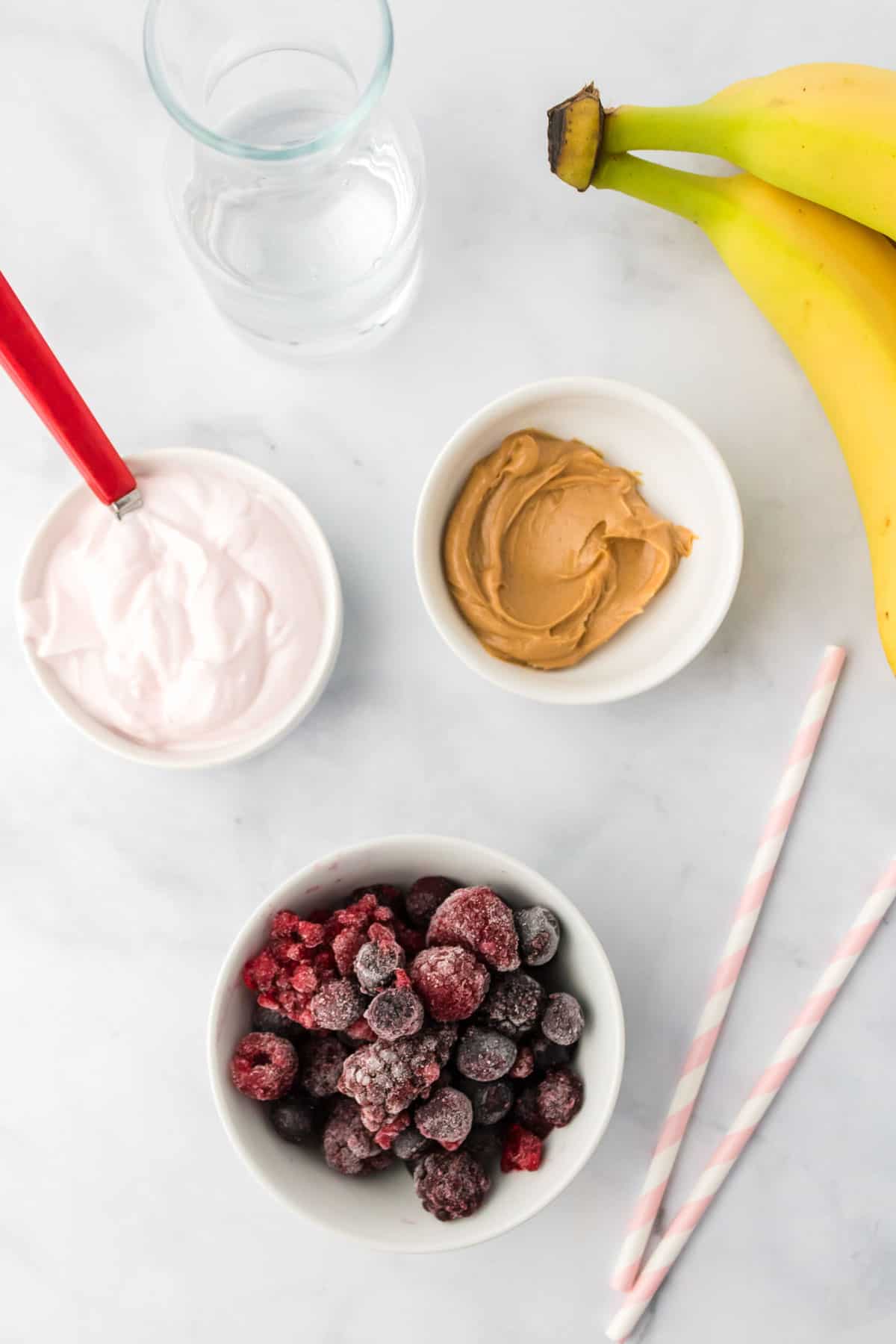 bowls of frozen berries, strawberry yogurt, peanut butter and water, a banana, and two striped straws on a marble tabletop.