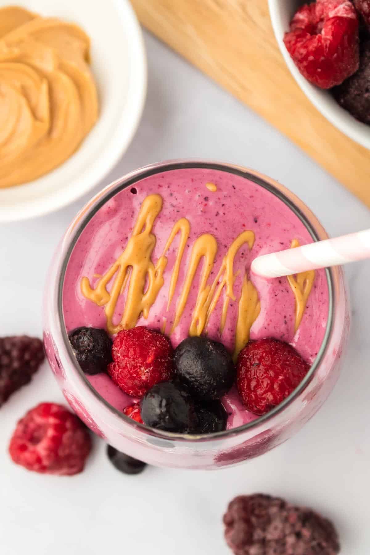 Looking down at a smoothie in a glass topped with a drizzle of peanut butter and mixed berries surrounded by a bowl of peanut butter and one of berries.