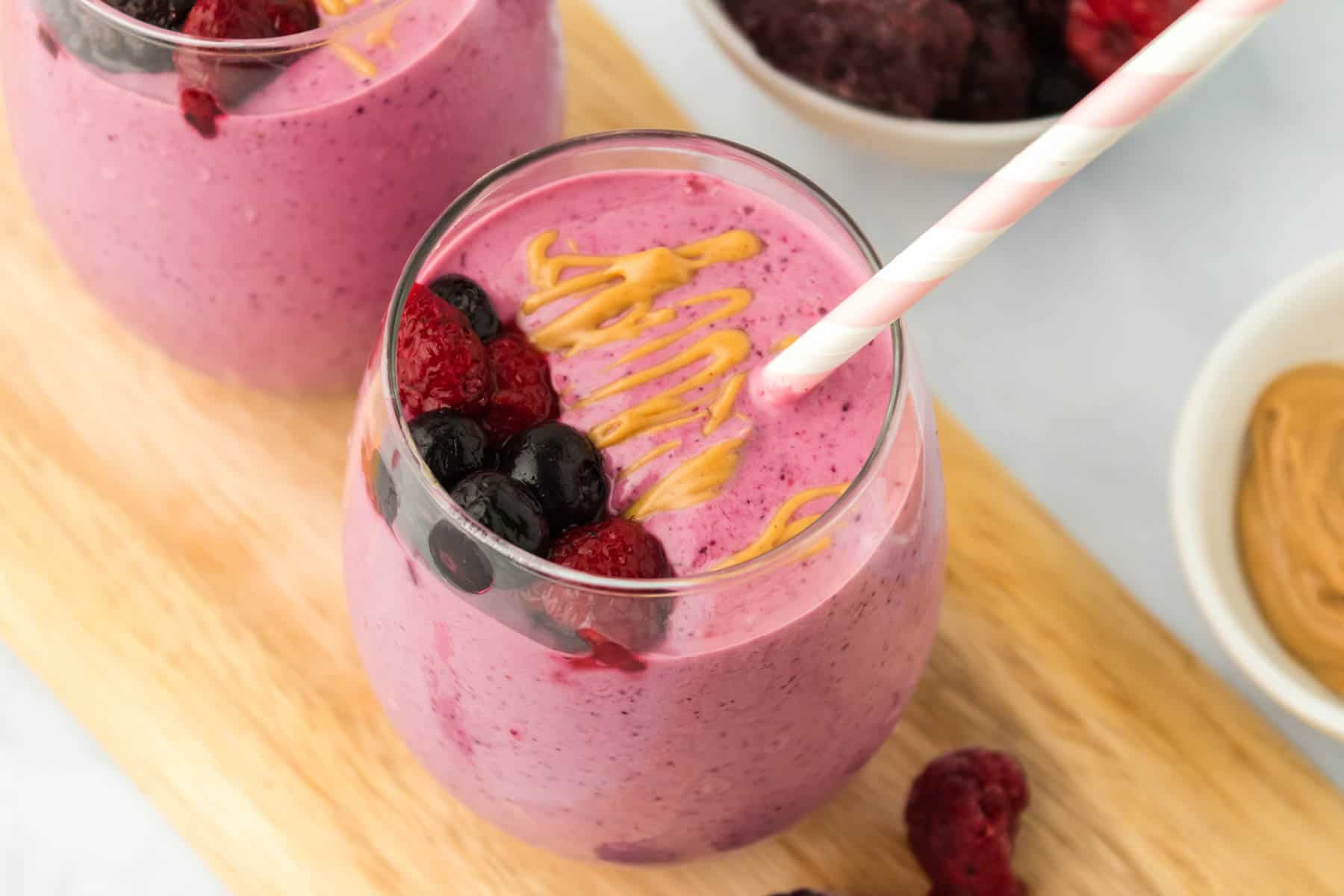 two peanut butter and jelly smoothies on a wooden board on a table with bowls of frozen mixed berries and peanut butter