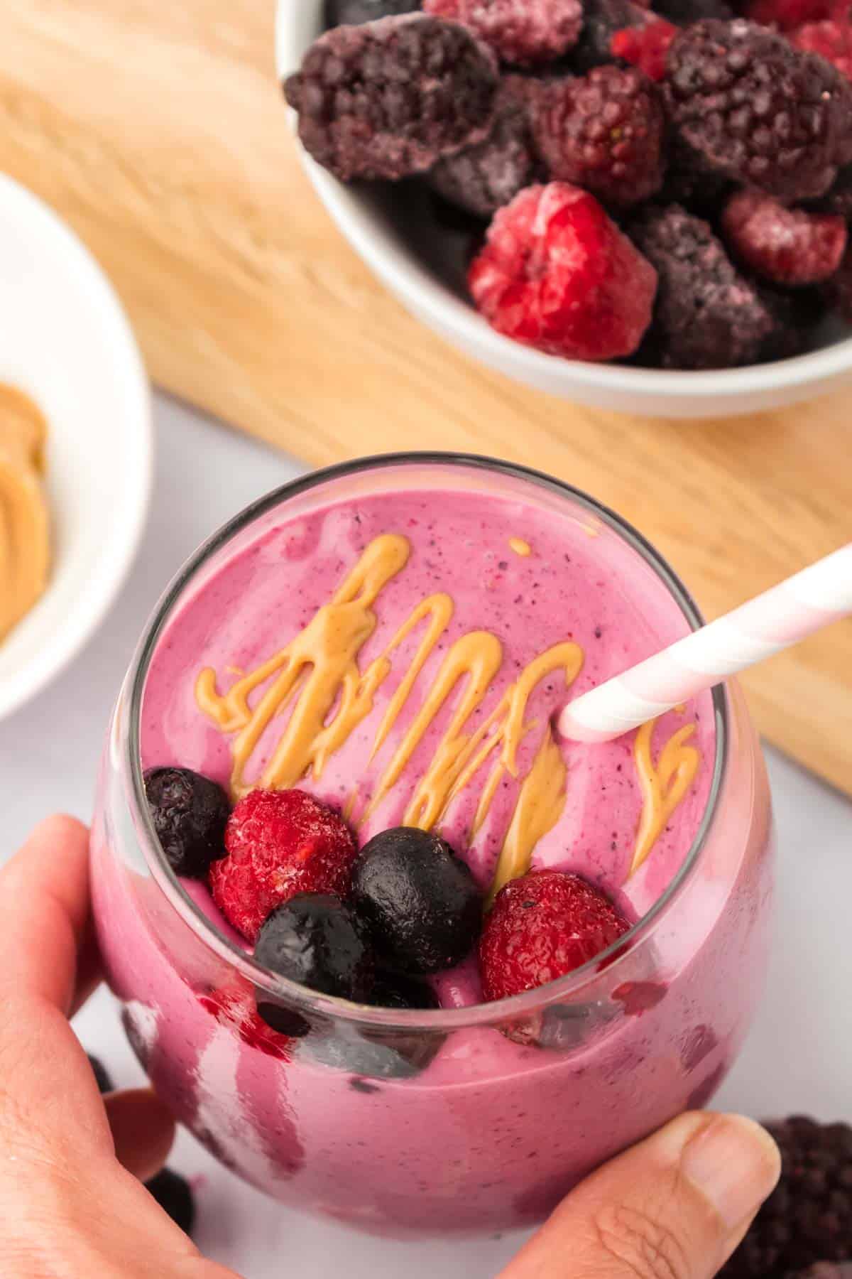 A hand holding a peanut butter and jelly smoothie in a glass next to a bowl of frozen berries.