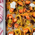 baked mini bell pepper nachos on a sheet pan with black beans, tomatoes, jalapenos, and melted cheese, and several topped with sour cream