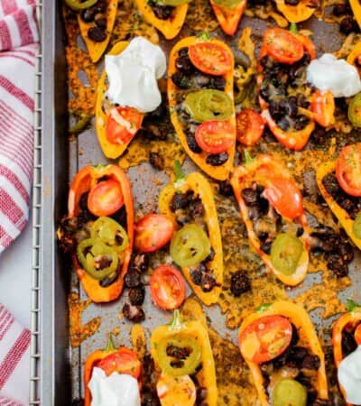 baked mini bell pepper nachos on a sheet pan with black beans, tomatoes, jalapenos, and melted cheese, and several topped with sour cream