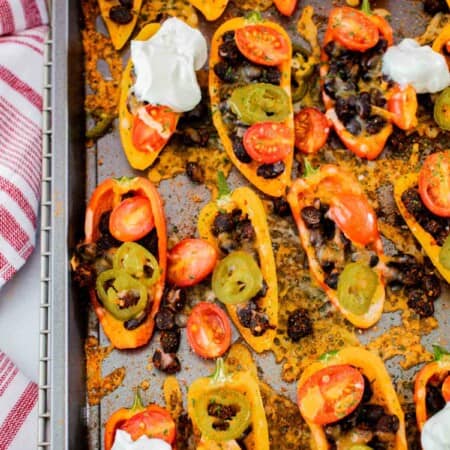 baked mini bell pepper nachos on a sheet pan with black beans, tomatoes, jalapenos, and melted cheese, and several topped with sour cream