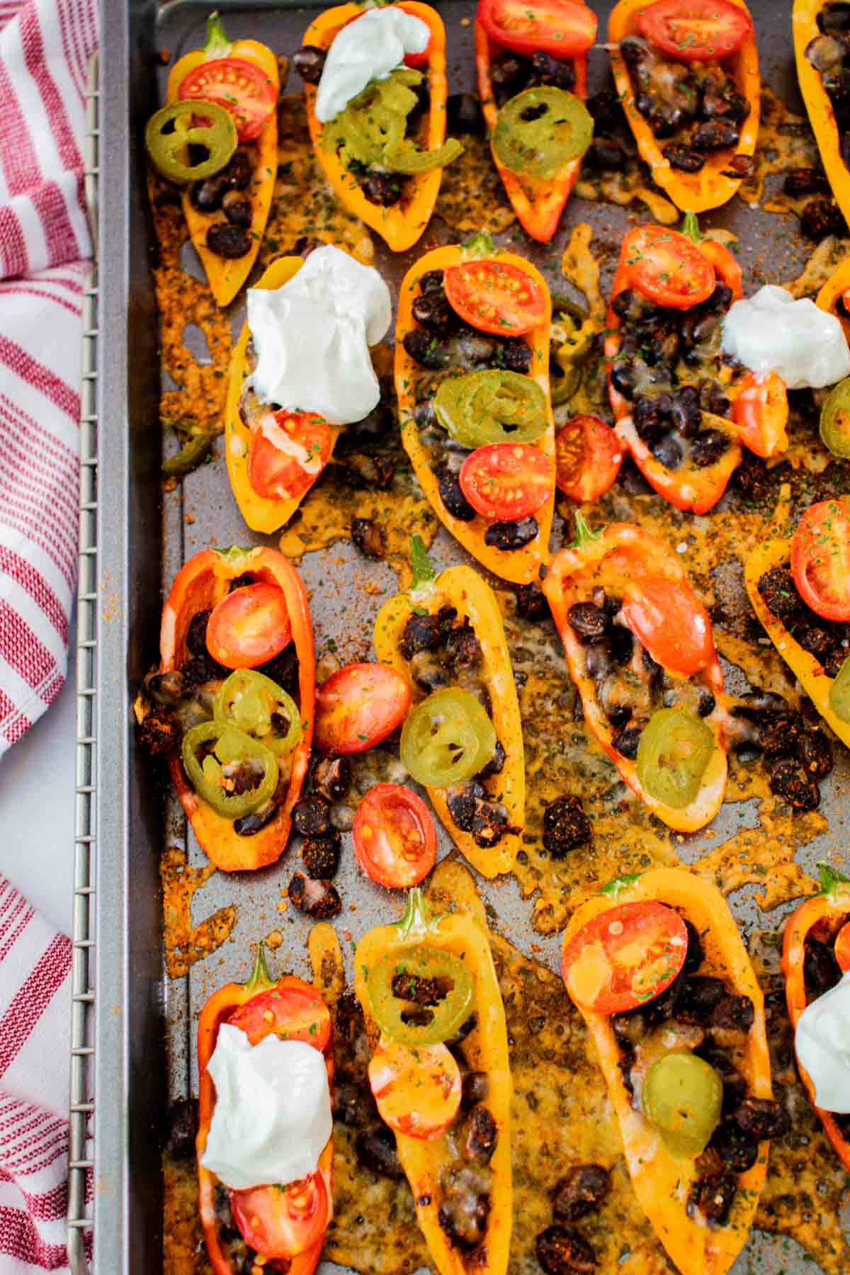 baked mini bell pepper nachos on a sheet pan with black beans, tomatoes, jalapenos, and melted cheese, and several topped with sour cream
