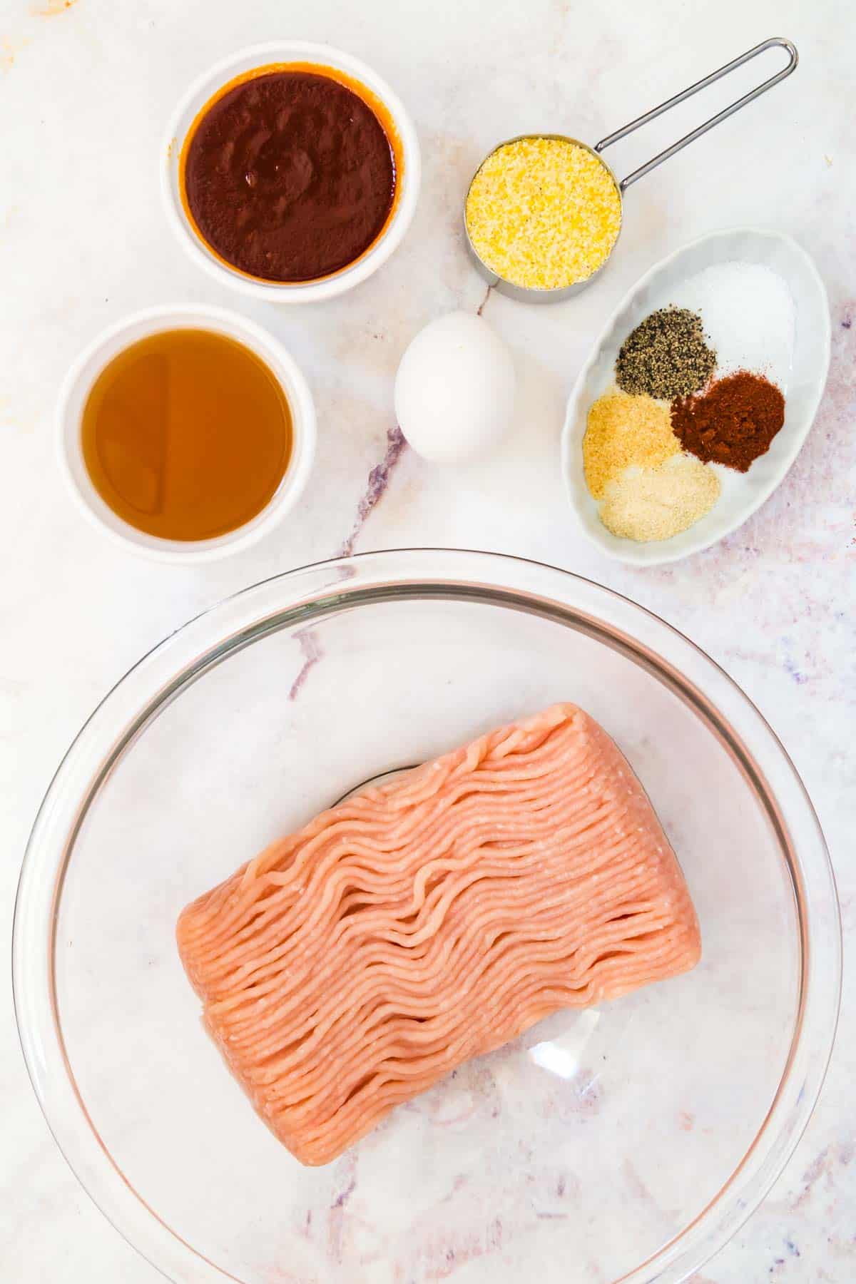 raw ground turkey in a bowl with bowls of barbecue sauce, apple juice, and spices, an egg, and a measuring cup with cornmeal