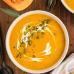 Overhead view of a bowl of butternut squash soup with cream, pepitas, and herbs on top.