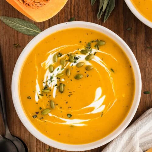 Overhead view of a bowl of butternut squash soup with cream, pepitas, and herbs on top.