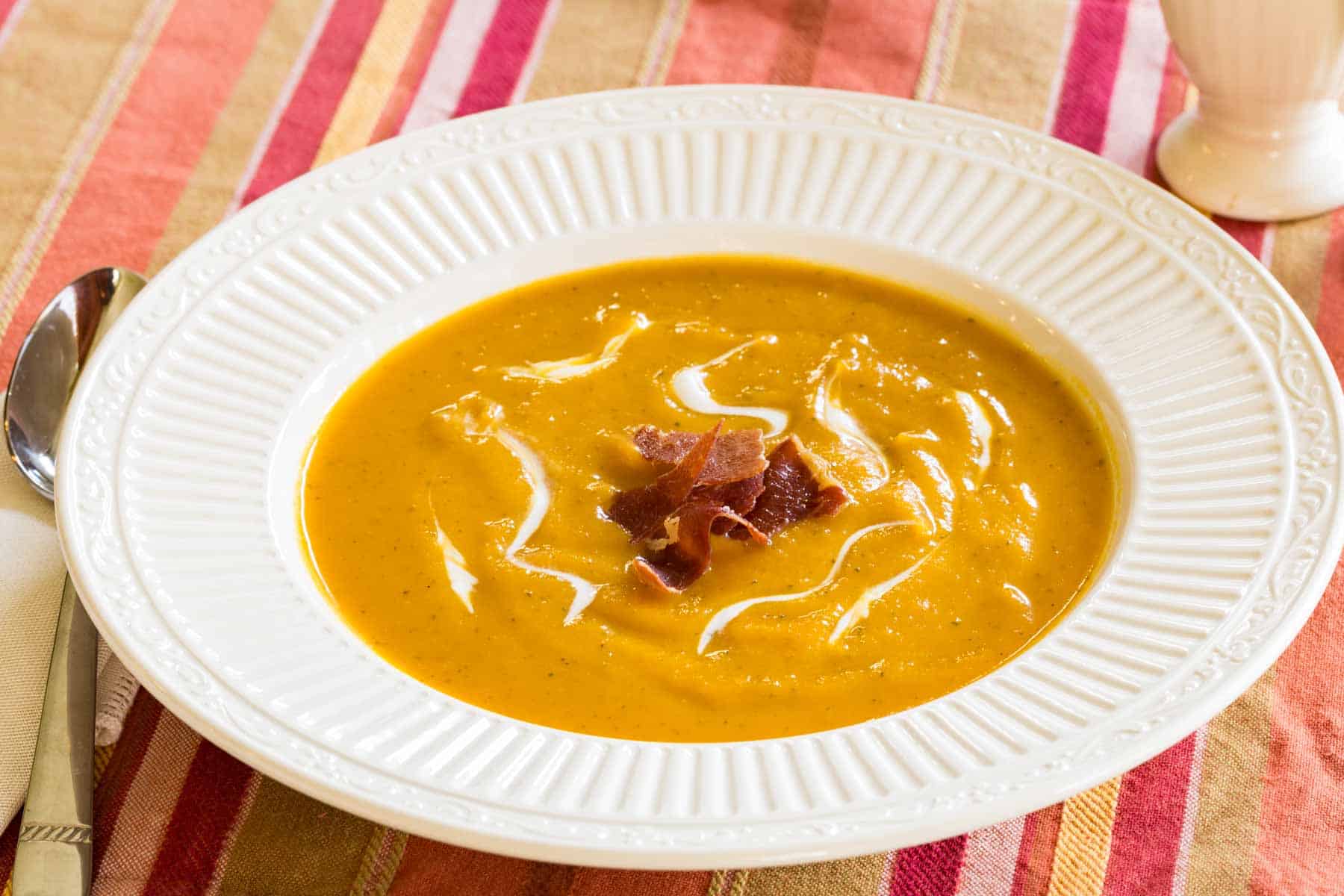 bowl of butternut squash soup garnished with crispy prosciutto next to a spoon and a cloth napkin on a striped placemat