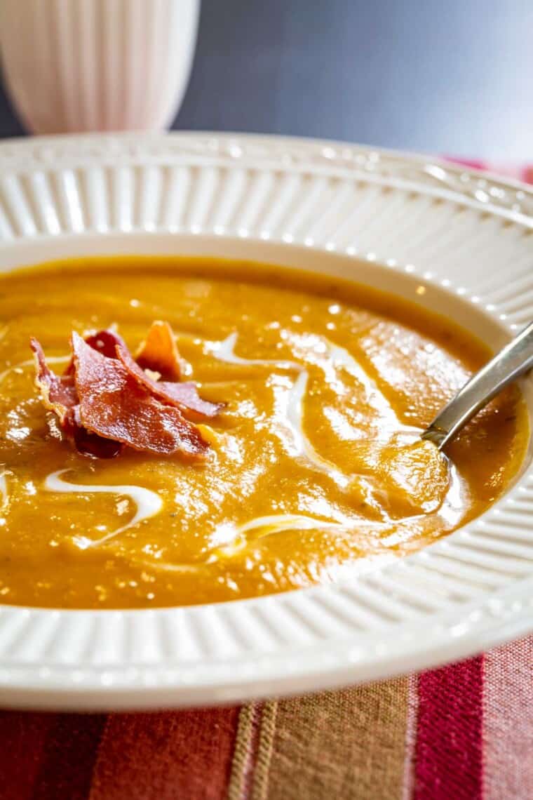 spoon set in a bowl of a creamy orange-colored pureed soup topped with crispy bits of prosciutto