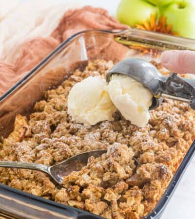 serving spoon resting in abaking dish of apple crisp with a scoops of ice cream being added on top