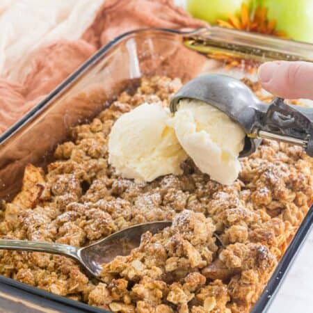 serving spoon resting in abaking dish of apple crisp with a scoops of ice cream being added on top