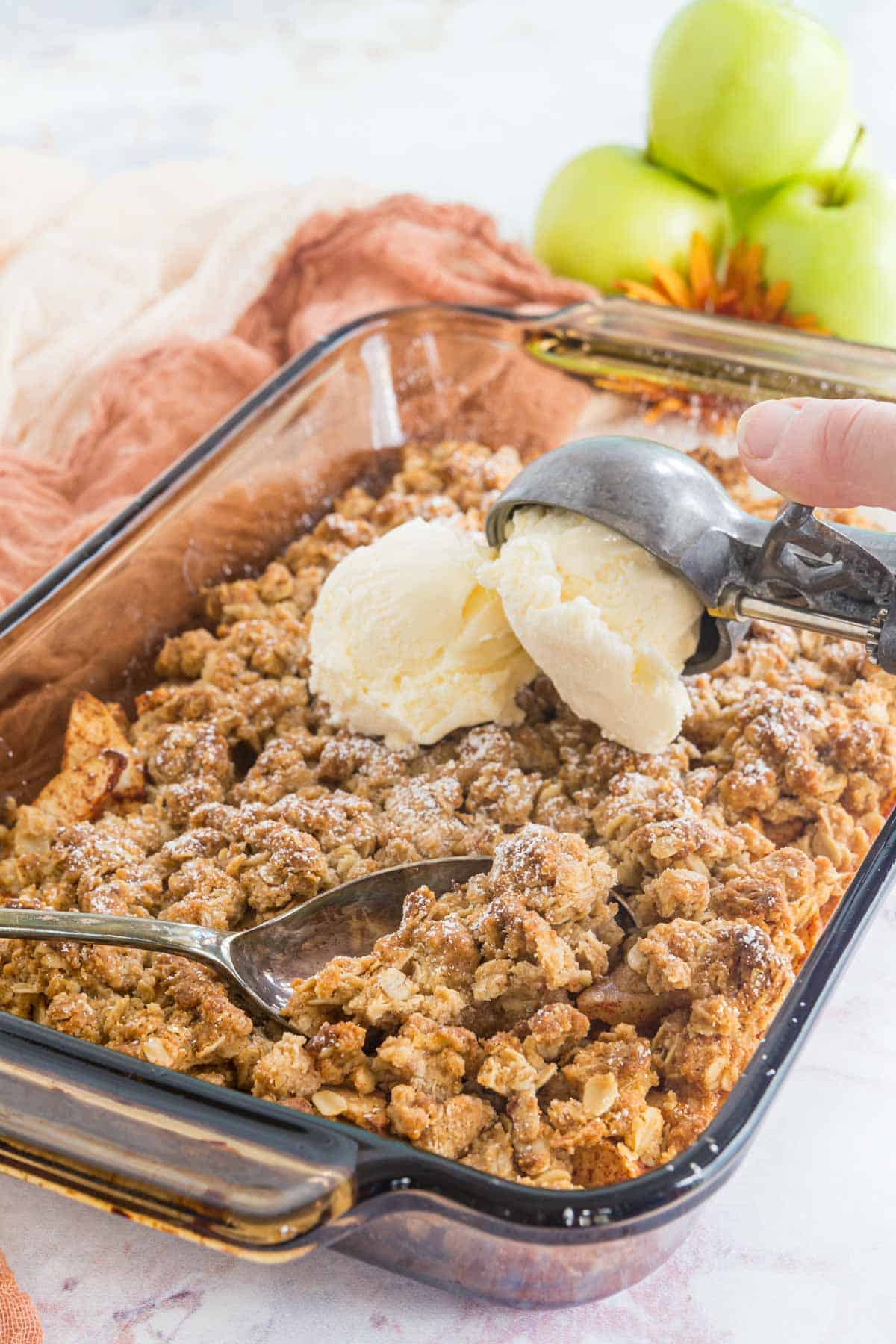 serving spoon resting in abaking dish of apple crisp with a scoops of ice cream being added on top