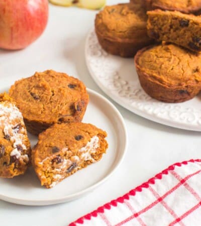 two apple pumpkin muffins with raisins on a plate with one cut in half and spread with butter next to a platter with more muffins