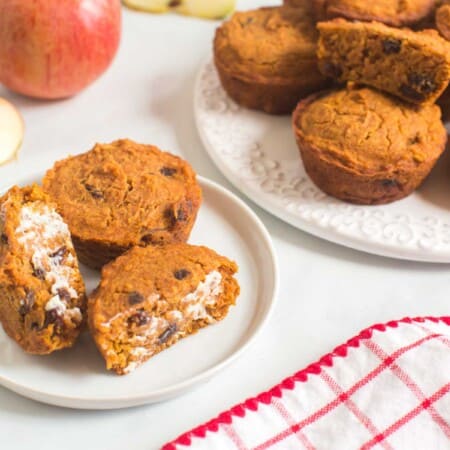 two apple pumpkin muffins with raisins on a plate with one cut in half and spread with butter next to a platter with more muffins