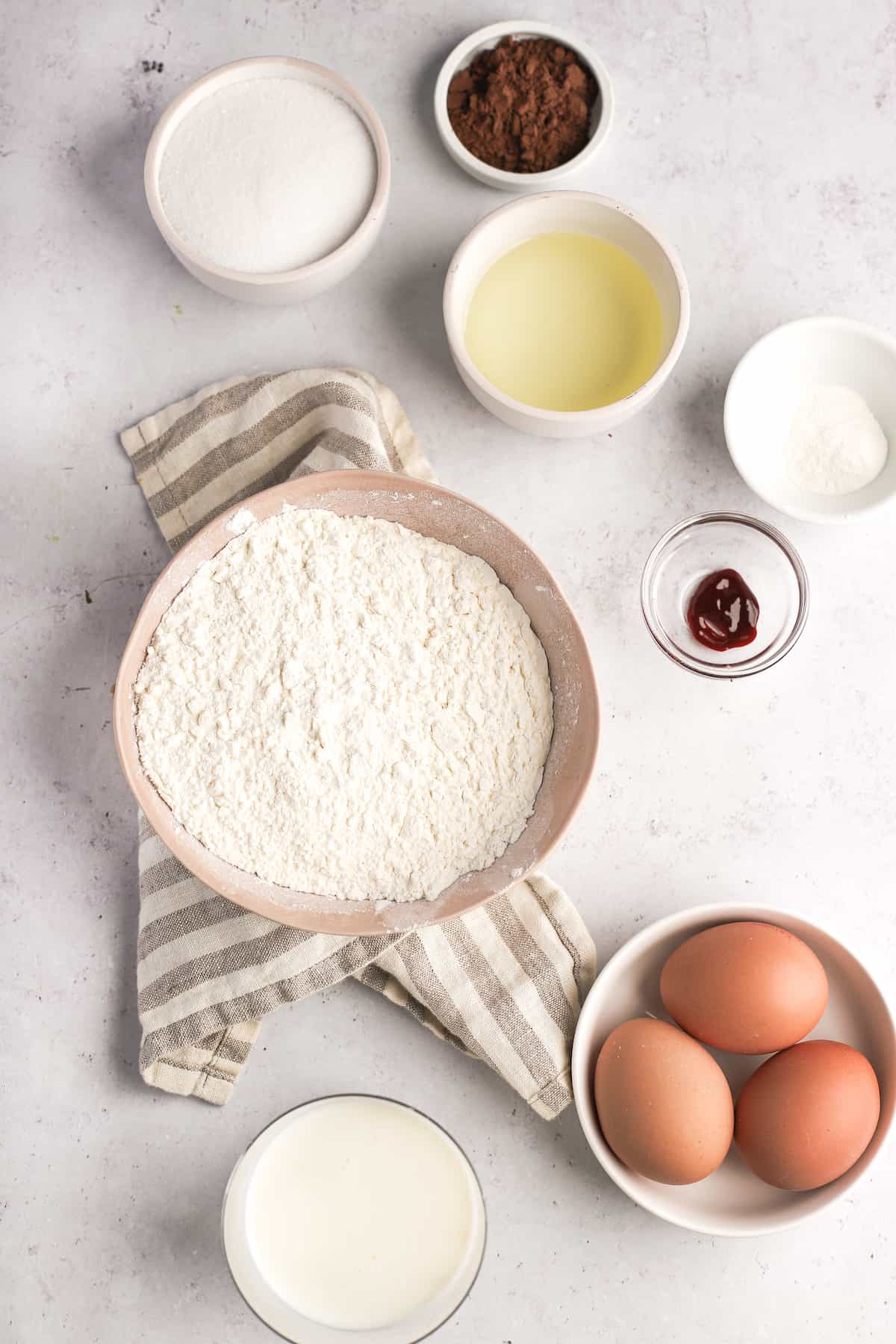 baking ingredients on a white background with flour, eggs, oil, food coloring and cocoa powder