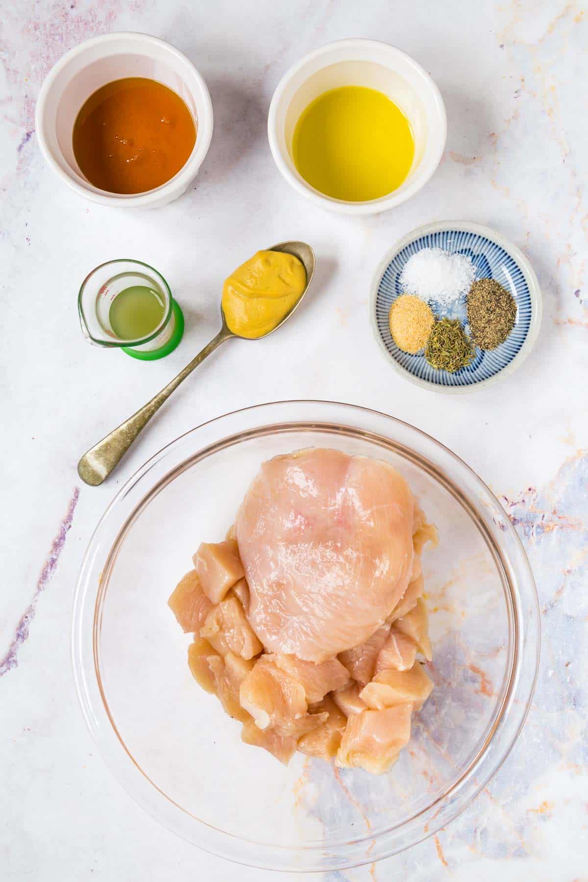 raw chicken in a bowl with some cut into cubes, a spoon of dijon mustard, bowls of honey, olive oil, salt, pepper, garlic powder, and thyme, and a small beaker with apple cider vinegar
