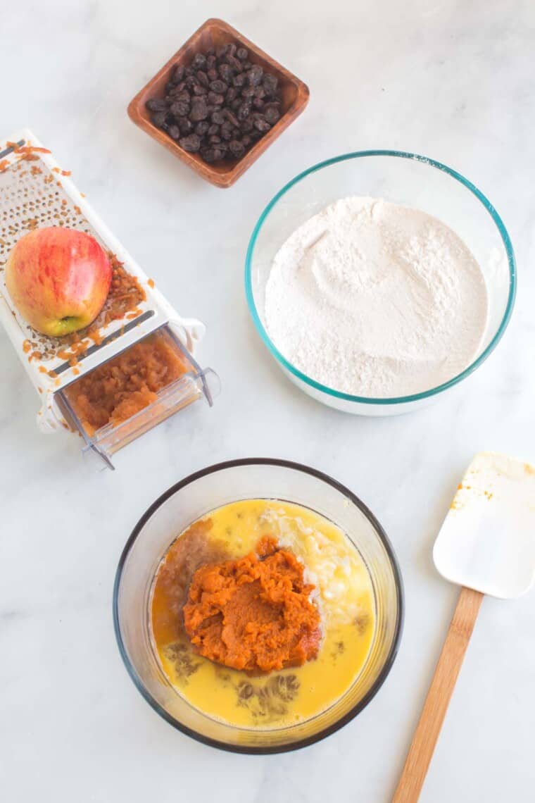 A bowl of the whisked dry ingredients, a bowl with the beaten egg, pumpkin puree, and other wet ingredients, a grater with grated apple, a bowl of raisins, and a spatula on a tabletop