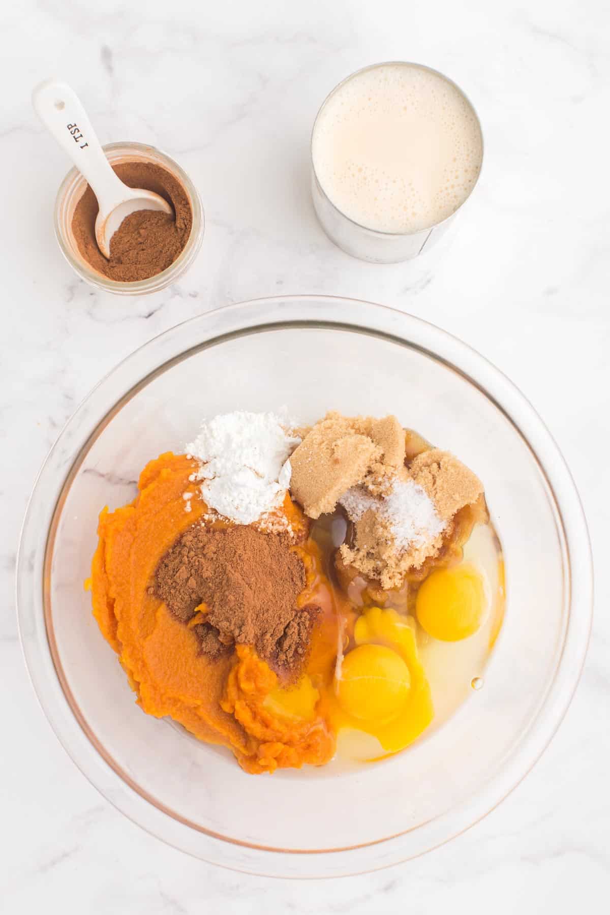 glass bowl with pumpkin puree, two eggs, light brow sugar, salt, cornstarch, and pumpkin pie spice on a marble counter top with a jar of pumpkin pie spice and a can of evaporated milk